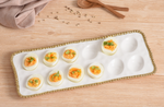 A top-down photograph of eight deviled eggs with vibrant orange filling and green herb garnish on a white porcelain platter with a gold beaded rim, arranged on a light wooden table with scattered white peppercorns