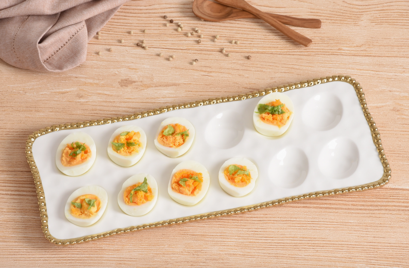 A top-down photograph of eight deviled eggs with vibrant orange filling and green herb garnish on a white porcelain platter with a gold beaded rim, arranged on a light wooden table with scattered white peppercorns