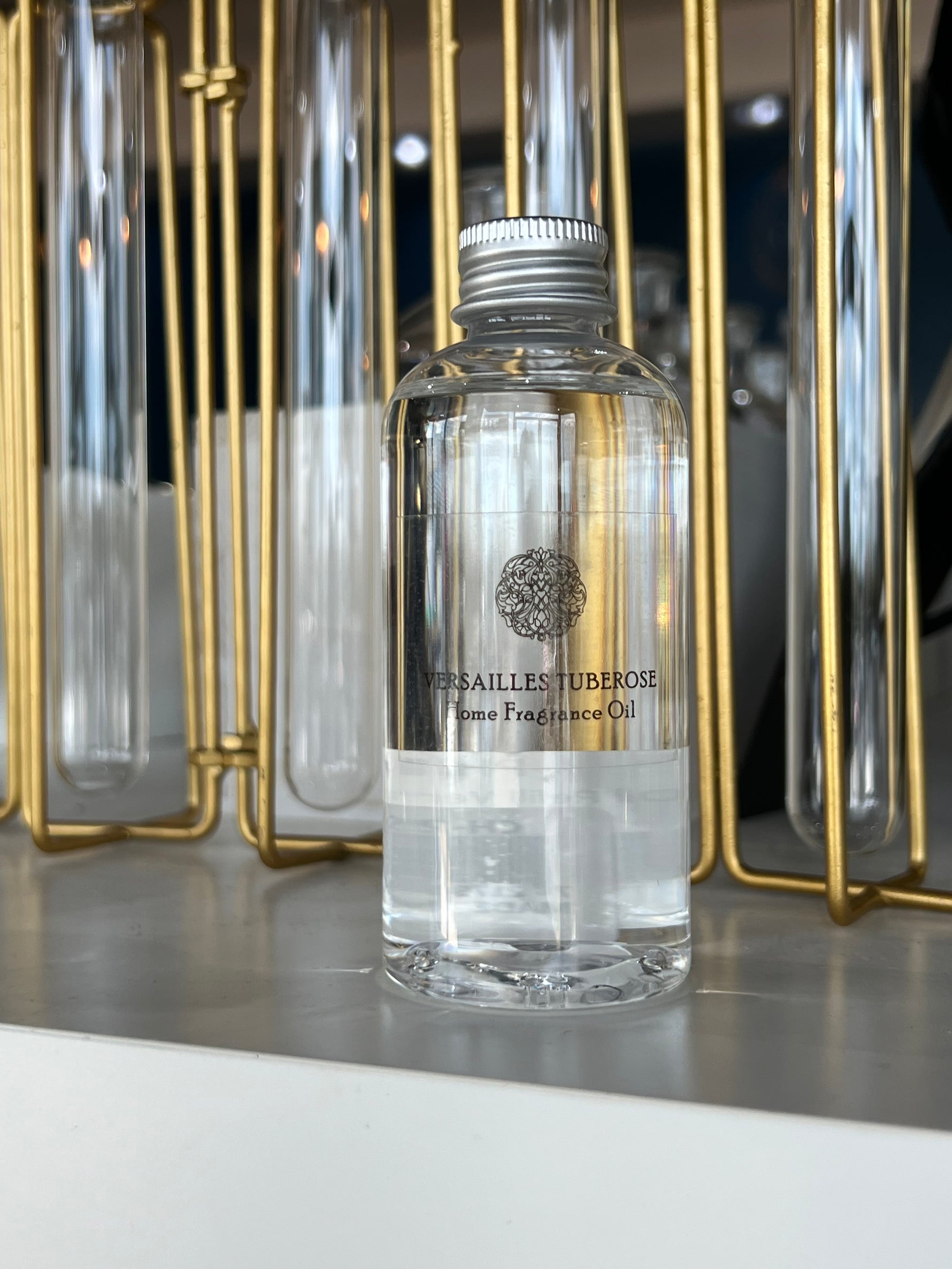 Photograph of a Versailles Tuberose Home Fragrance Oil bottle featuring an ornate black filigree logo and silver ribbed cap set against a backdrop of gold-colored metal rods and clear glass cylinders