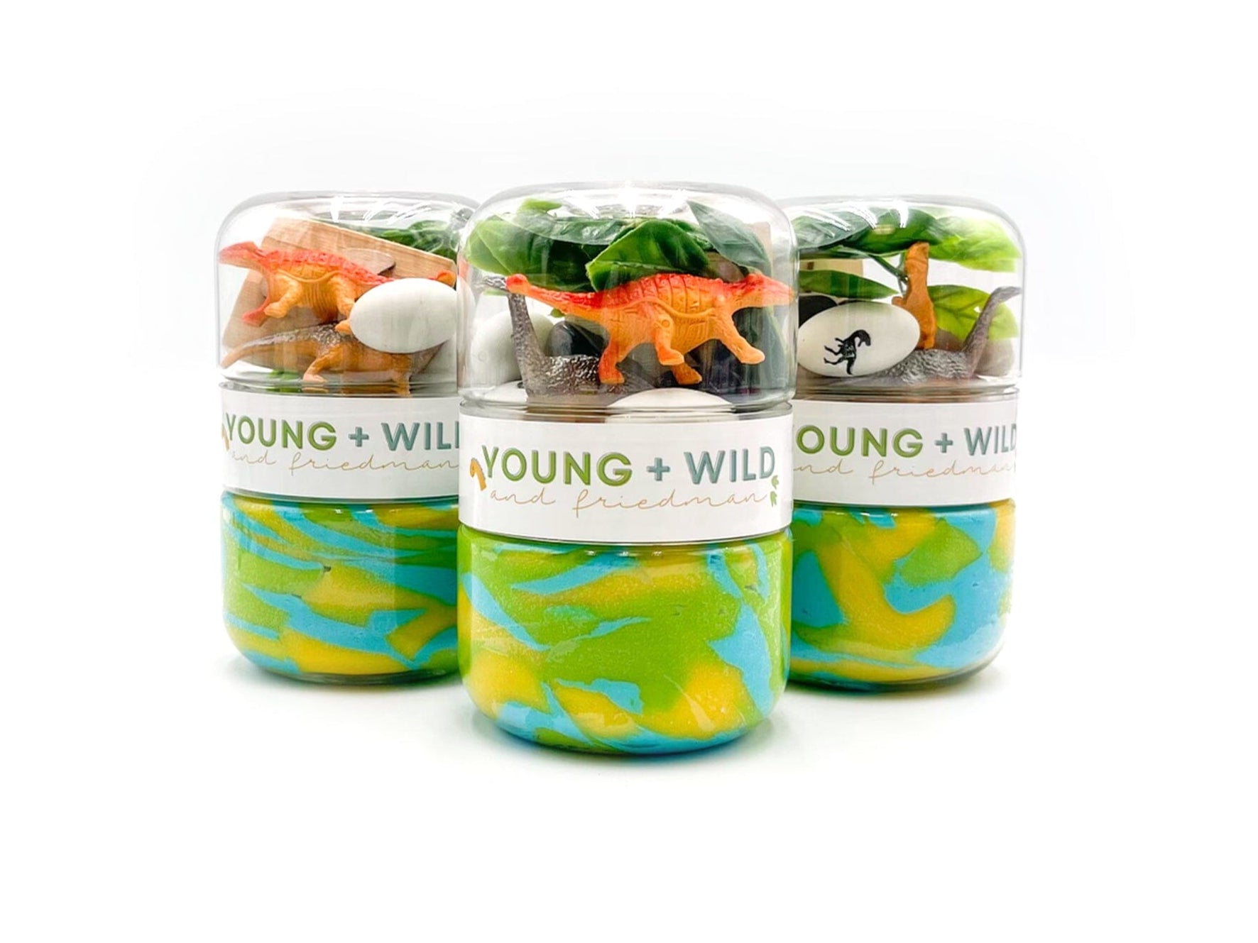 Young + Wild and Friedman's dinosaur-themed playdough kits feature three clear containers stacked, each displaying small orange and grey dinosaur toys, white eggs, and green foliage over vibrant swirled blue, green, and yellow playdough on a clean white background