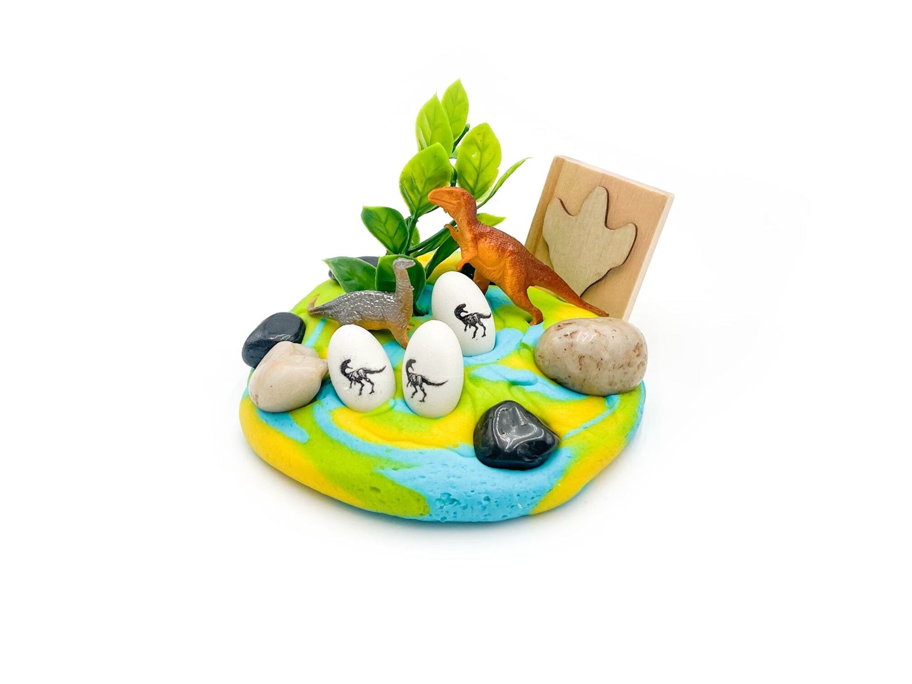 A vibrant miniature dinosaur play scene with an orange T-Rex toy, a grey Brachiosaurus toy, three white dinosaur eggs with black skeleton decals, green foliage, and a wooden dinosaur footprint on a marbled blue, yellow, and green base