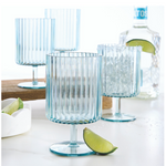 Four light blue ribbed stemmed glasses, one with a salt-rim and ice, are arranged on a white marble surface with fresh lime wedges and a bottle of Altos tequila in the background