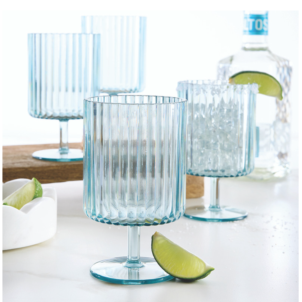 Four light blue ribbed stemmed glasses, one with a salt-rim and ice, are arranged on a white marble surface with fresh lime wedges and a bottle of Altos tequila in the background
