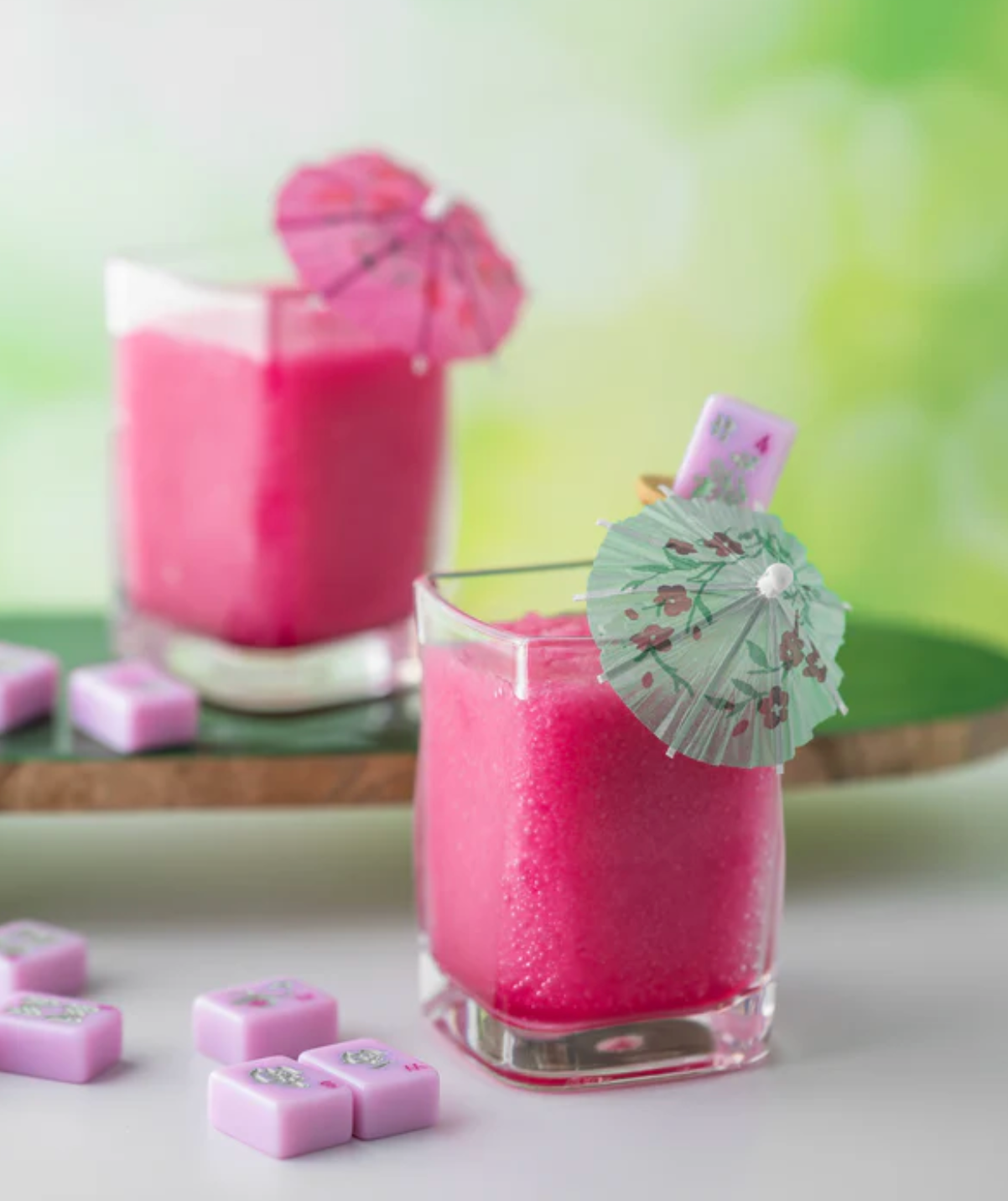 Two glasses of pink slushy drinks with cocktail umbrellas and mahjong tiles on a white surface