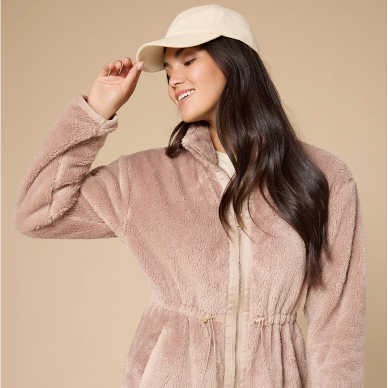 A smiling young woman wearing a cream baseball cap and a plush dusty rose fleece jacket with a gold zipper and drawstring waist on a plain background