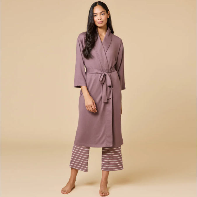 A woman with long dark hair models a long-sleeved mauve robe tied at the waist over white horizontal striped capri pants, standing barefoot against a plain beige studio background