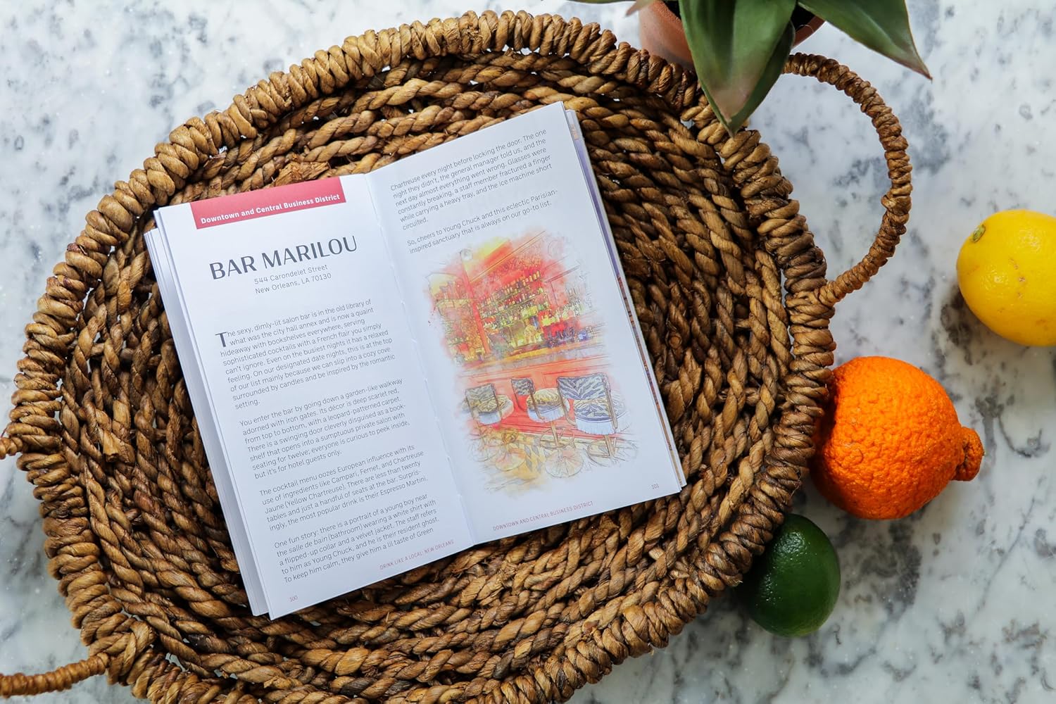 Open magazine page with an illustrated article on Bar Marilou in New Orleans, LA, nestled in a round woven basket on white marble with a vivid yellow lemon, a bumpy orange tangerine, and a green lime