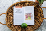An illustration from a publication depicts Kermit's Treme Mother In Law Lounge in New Orleans, Louisiana, resting open on a woven wicker basket with a fresh green lime on the page, featuring colorful murals with caricature faces on the building exterior