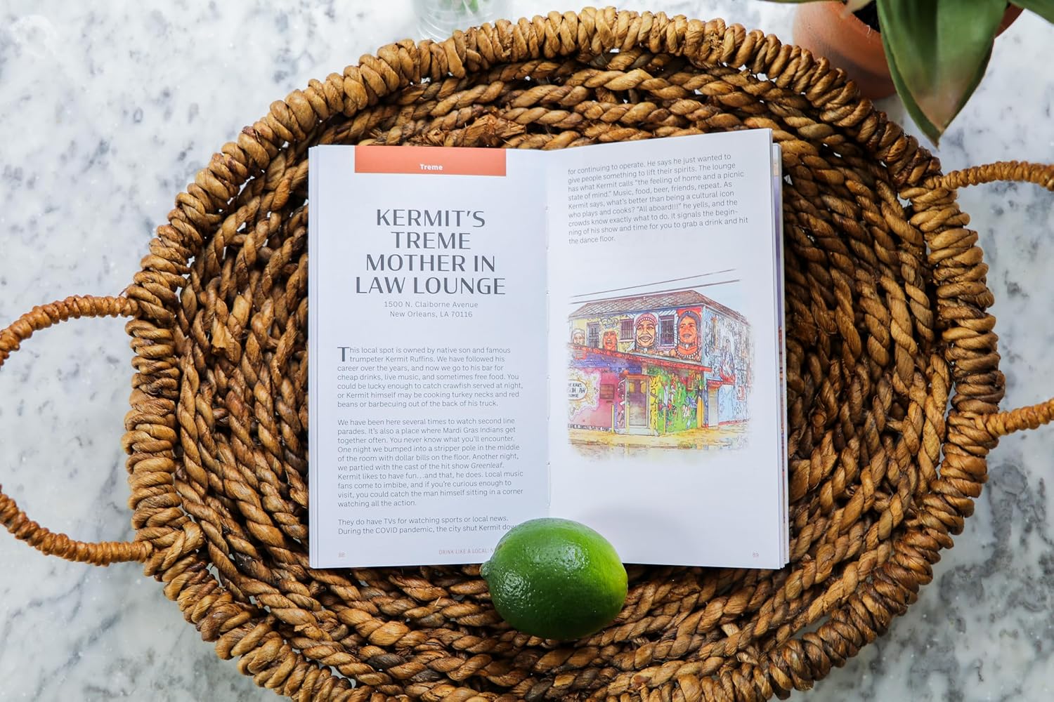 An illustration from a publication depicts Kermit's Treme Mother In Law Lounge in New Orleans, Louisiana, resting open on a woven wicker basket with a fresh green lime on the page, featuring colorful murals with caricature faces on the building exterior