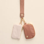 Two small pouches, one light pink and one dusty rose with subtle vertical ribbing, featuring gold-toned zippers and clasps, hanging from a matching woven pink strap against a light background