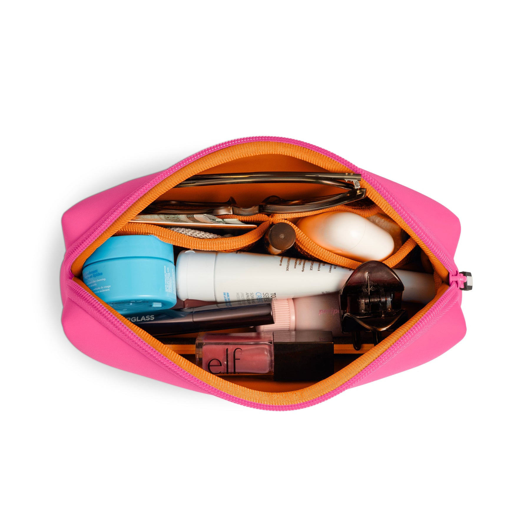 A vibrant fuchsia pink and orange zippered makeup bag on a white background, filled with various beauty essentials including a light blue skincare jar, a white TOPI CREM tube, pink elf lip gloss, sunglasses, and a dark hair claw clip