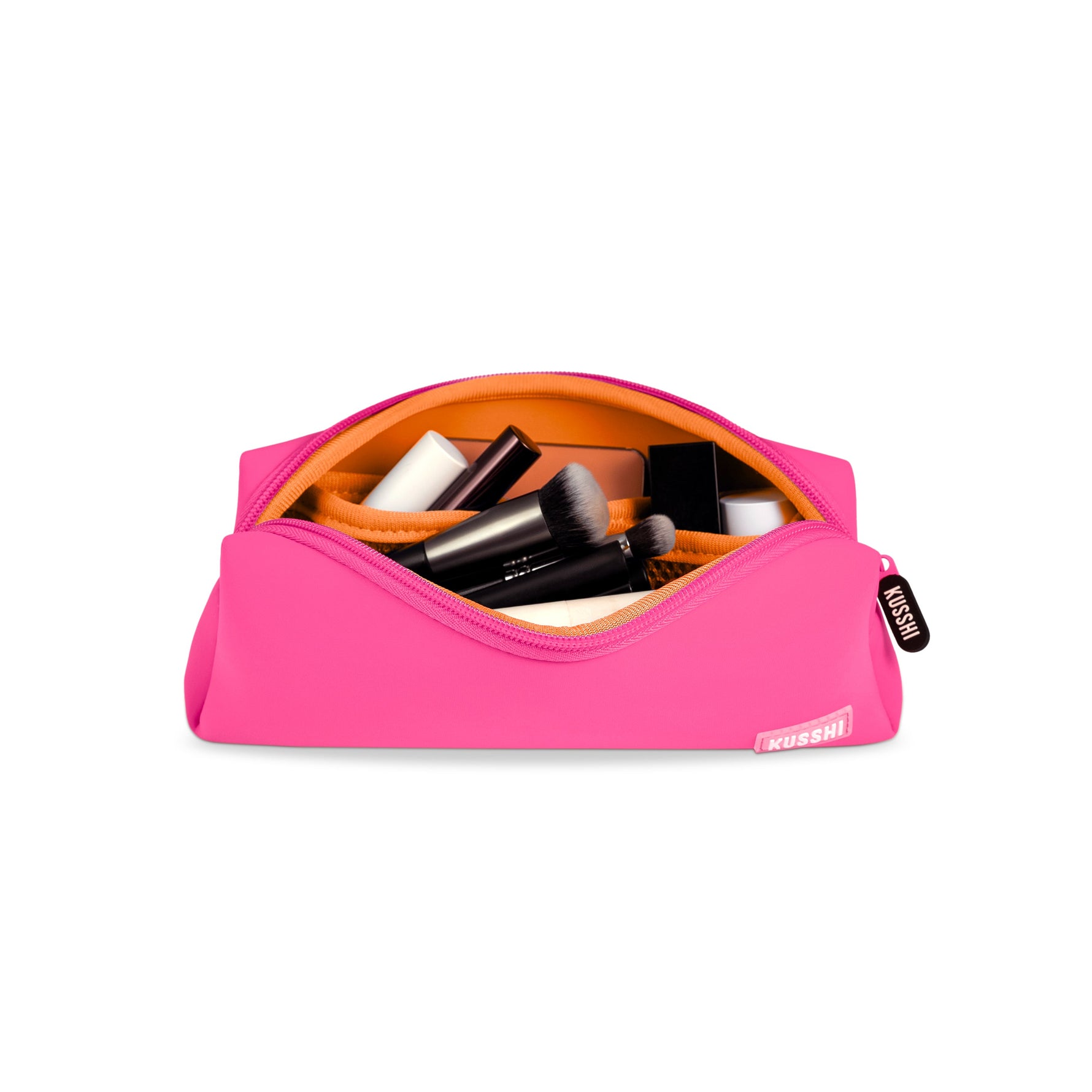 A hot pink Kusshi makeup bag with a bright orange interior filled with several black makeup brushes and other cosmetic items on a white background