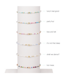 Product photography of six delicate beaded bracelets, each featuring tiny colorful beads and a central gold bead, displayed vertically on a white stand with distinct styles like turquoise, pastel multi-color, and a vibrant rainbow design, each with an identifying text label such as turq it real good and hot mess