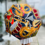 An artistic umbrella with a colorful pattern of gold trumpets, red drums, and blue saxophones is held by a person in a white pleated skirt on an outdoor patio