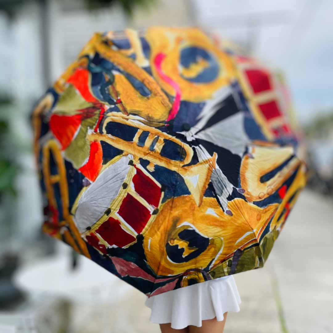 An artistic umbrella with a colorful pattern of gold trumpets, red drums, and blue saxophones is held by a person in a white pleated skirt on an outdoor patio