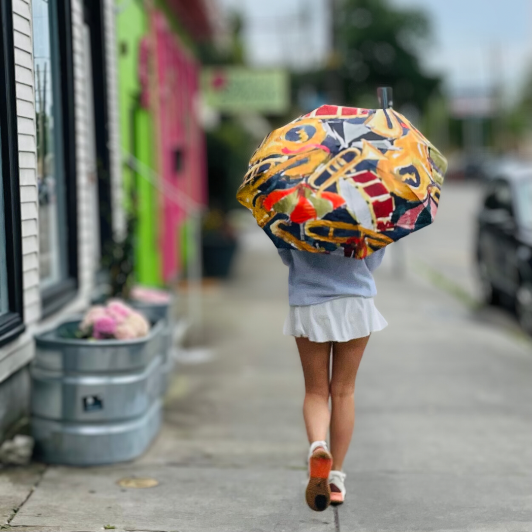 A person walks away on a city sidewalk holding a vibrant yellow, red, and blue musical instrument patterned umbrella, passing brightly colored pink and lime green buildings and silver planters