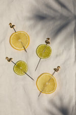 Photograph of four vibrant lemon and lime slices with golden flamingo cocktail picks on a white linen surface under palm leaf shadows
