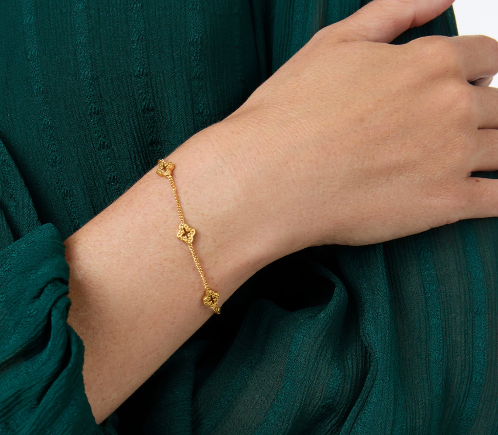 Gold chain bracelet with three clover charms worn on a wrist against a green fabric background