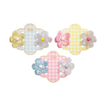 Mudpie resin hair clips, three sets of flower-shaped clear pearlescent clips featuring colorful yellow, pink, and blue centers, displayed on sweet gingham backing cards in coordinating pastel blue, yellow, and pink patterns