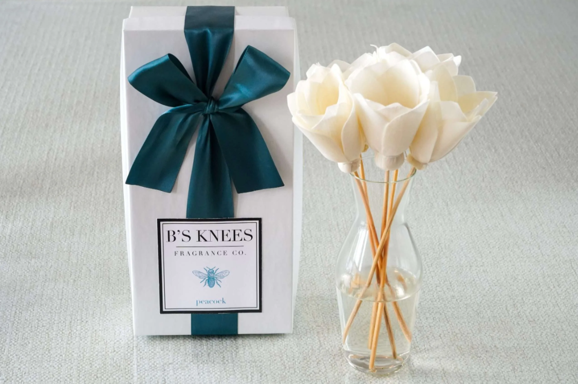 White gift box with teal ribbon next to a glass vase containing white flower reed diffusers