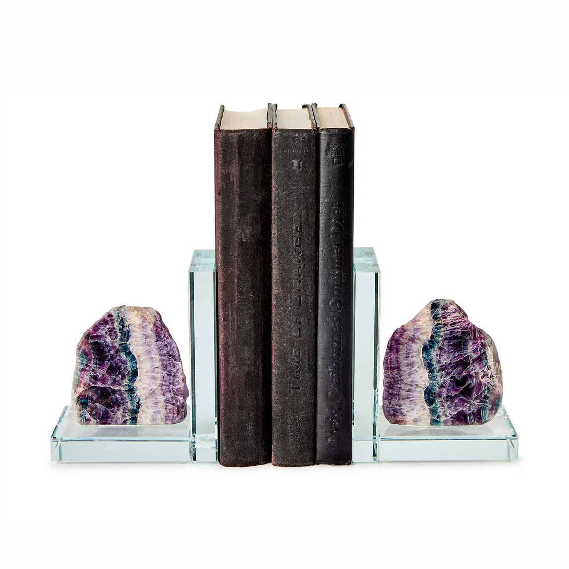 A studio photograph featuring a pair of raw fluorite crystal bookends with vibrant purple, teal, and white layers, holding three dark vintage books on clear glass bases