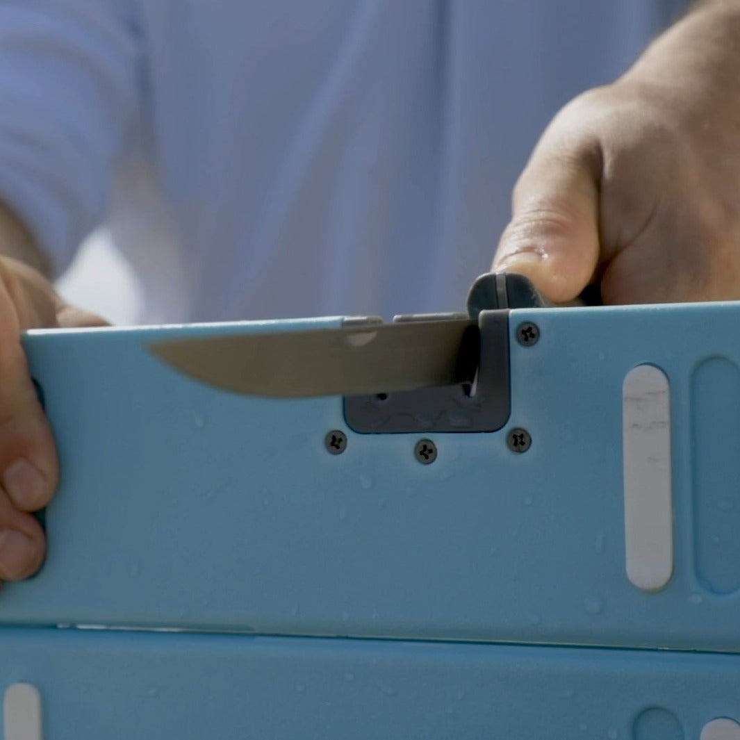 Photograph of a person's hands sharpening a silver-bladed knife in a gray slot on a light blue device covered in water droplets