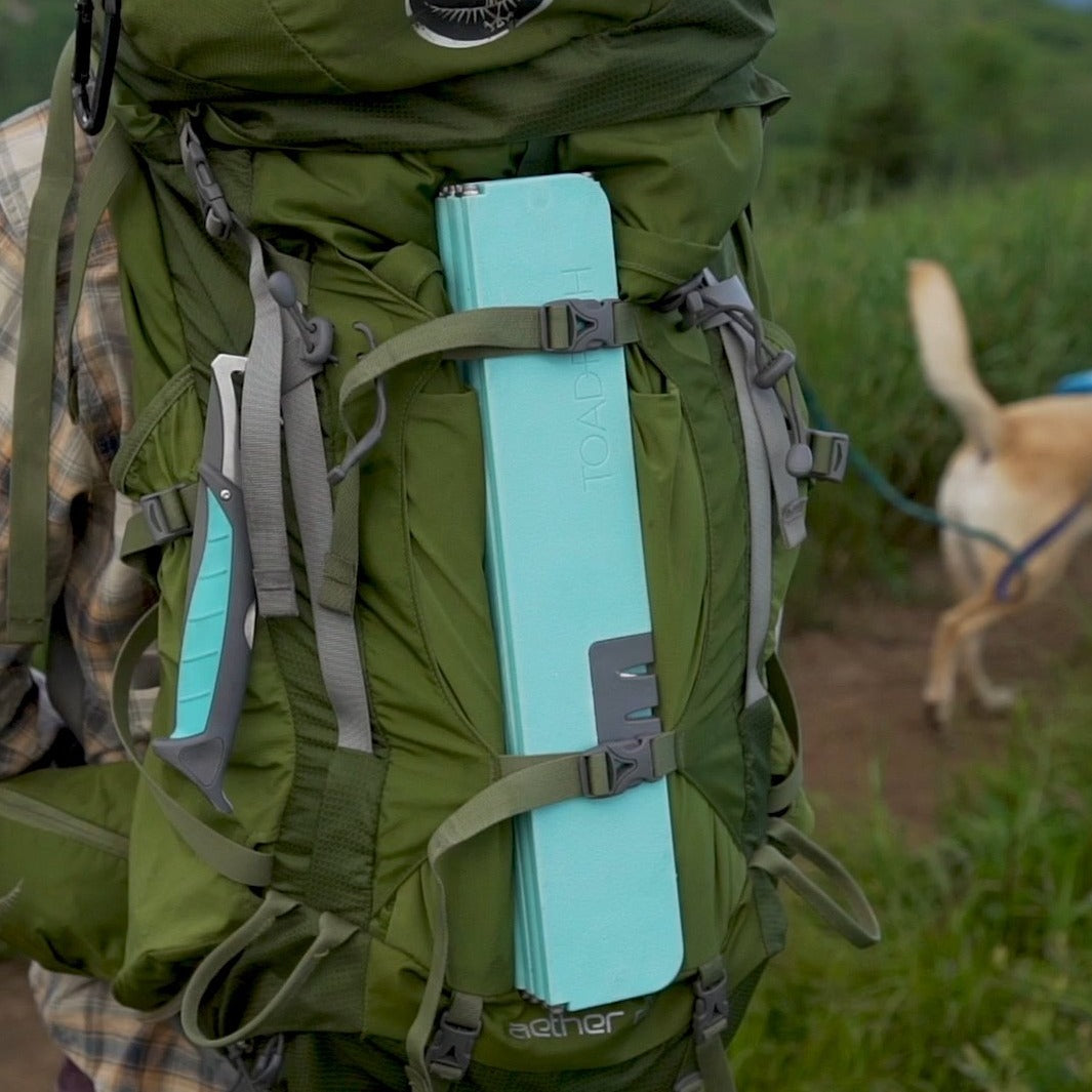 A green Osprey Aether hiking backpack on a dirt trail showing a light blue TOAD&CO foam pad strapped on one side and a turquoise-handled tool on the other, with a blurry yellow dog on a leash in the background