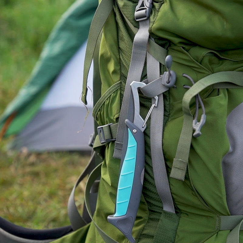 Photograph of a teal and grey folding camp saw clipped to an olive green hiking backpack at a grassy campsite with a tent in the background featuring a turquoise textured grip and grey nylon webbing straps