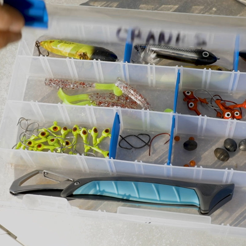 An open fishing tackle box displaying a yellow and black hard lure, clear glitter soft plastic worms with chartreuse tails, and numerous bright green jig heads with red eyes