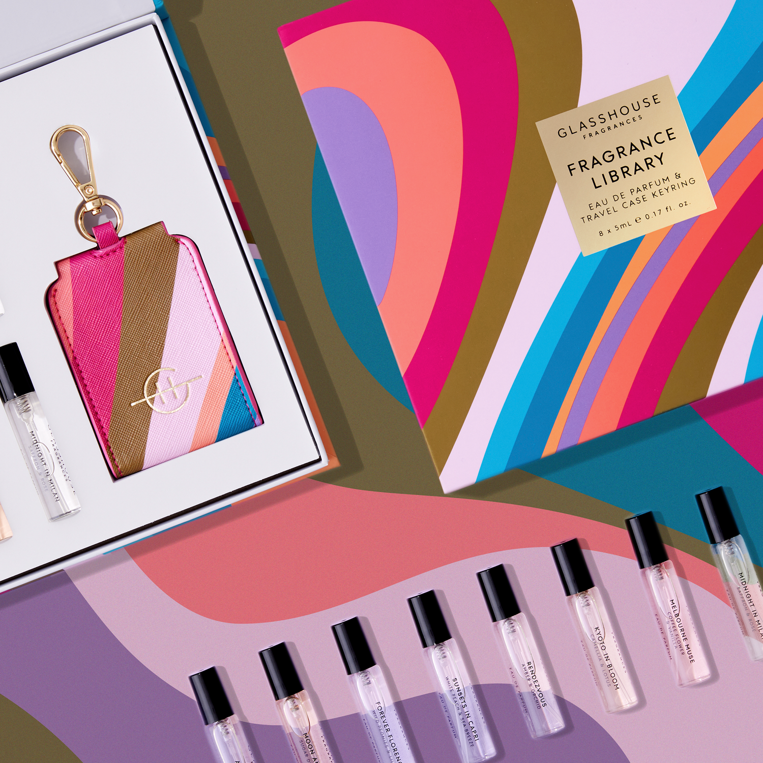 Glasshouse Fragrances Fragrance Library gift set displaying a gold-labeled box, a vibrant pink and gold striped travel case keyring, and multiple mini perfume vials like Midnight in Milan and Kyoto in Bloom, all presented on a colorful geometric background