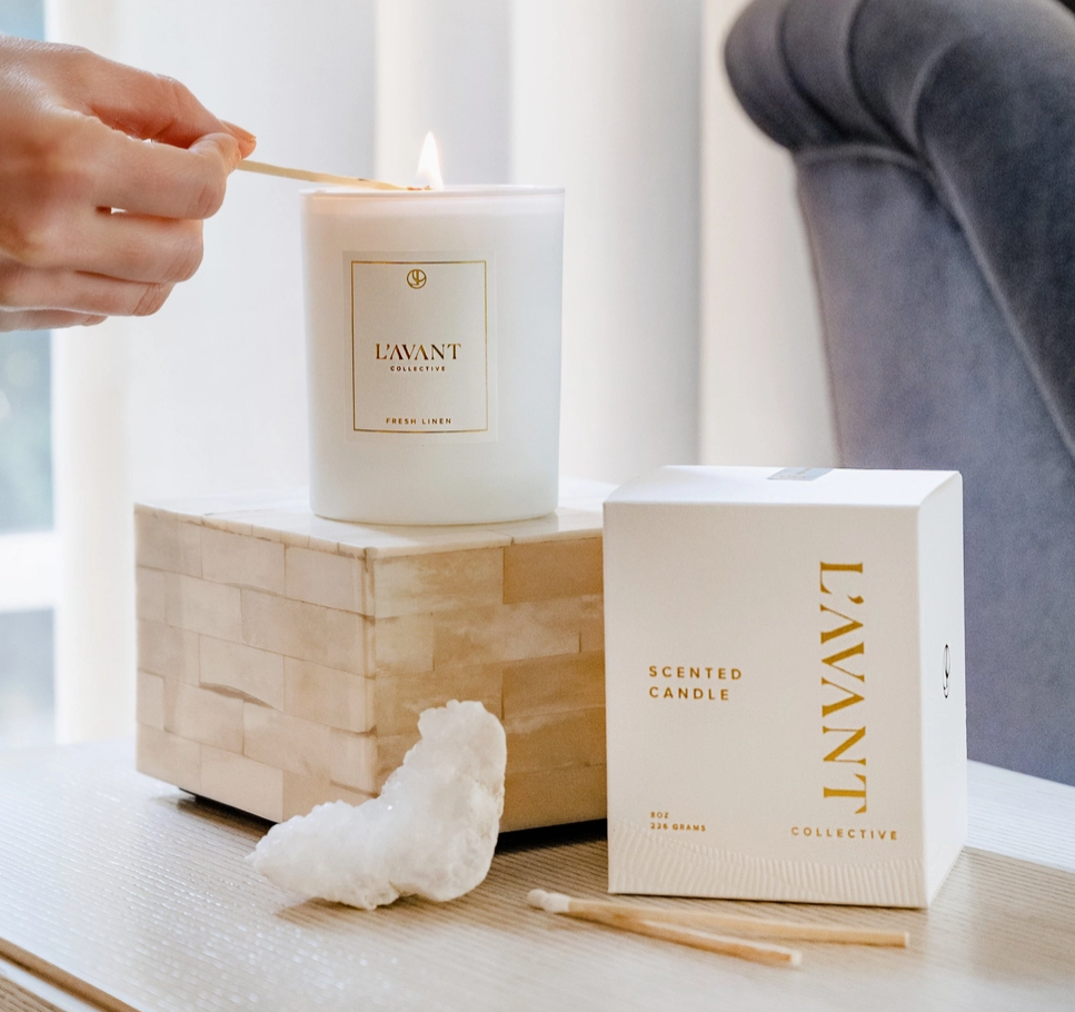 A hand lights a L'AVANT Collective Fresh Linen scented candle in a frosted white glass jar with a gold label, displayed on a mother-of-pearl tiled pedestal beside its matching box, a decorative white crystal, and wooden matchsticks