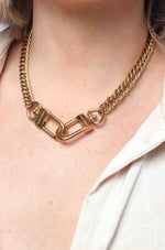 Photograph: A Marin Costello gold chunky chain necklace on a woman's décolletage, featuring two industrial carabiner-style links engraved with the brand name, worn over a cream collared shirt