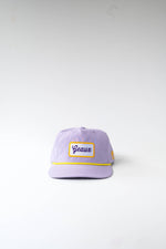 Photograph of a light purple baseball cap with a yellow rope accent and a white rectangular patch featuring "Geaux" embroidered in purple script and a yellow border, displayed on a plain white studio background
