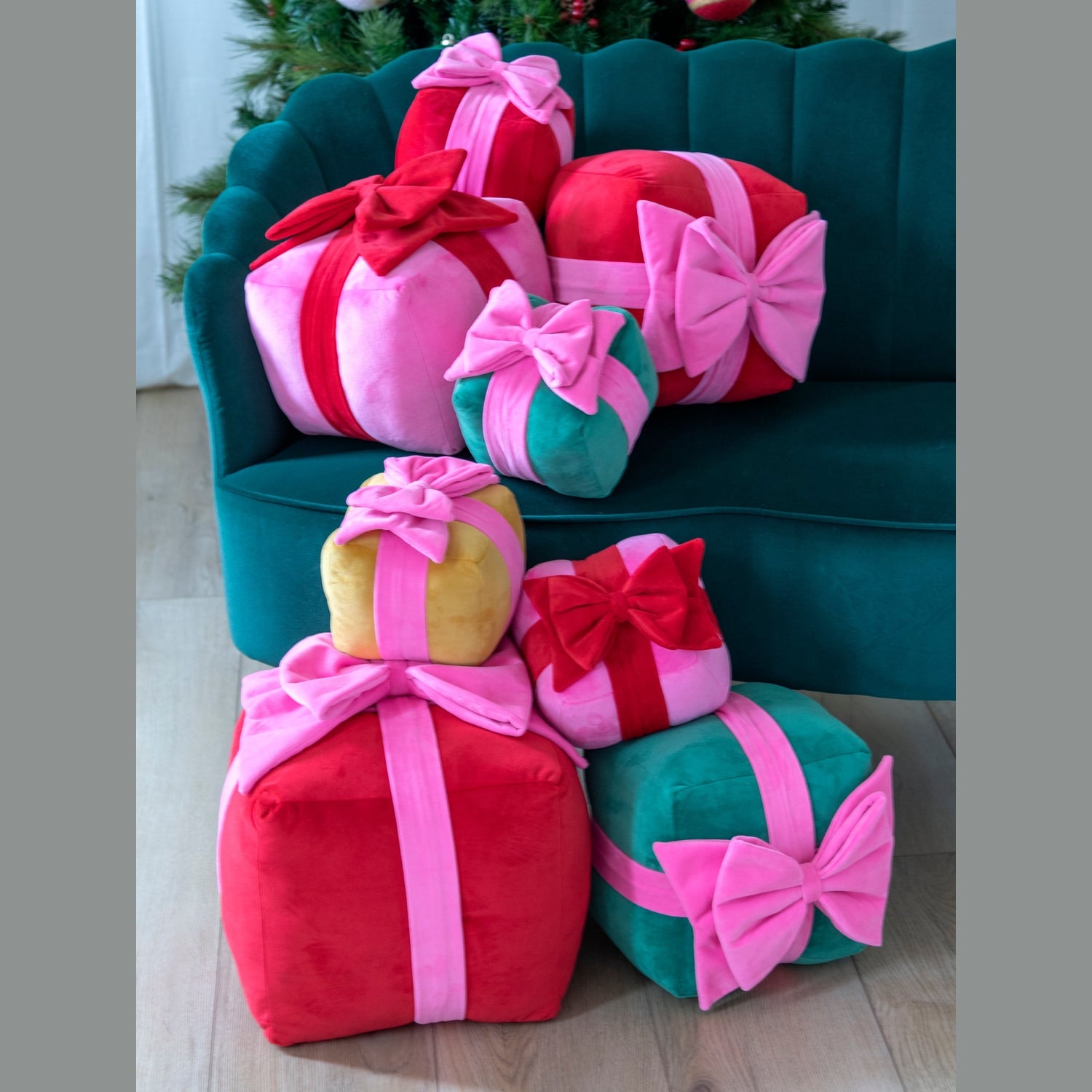 Stacked red, pink, green, and gold velvet gift-shaped pillows with large decorative bows on and beside a teal green scalloped sofa with a subtle Christmas tree in the background
