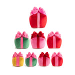 Studio shot of seven soft velvet gift box pillows in red, green, pink, and yellow, each tied with contrasting pink or red bows against a white background