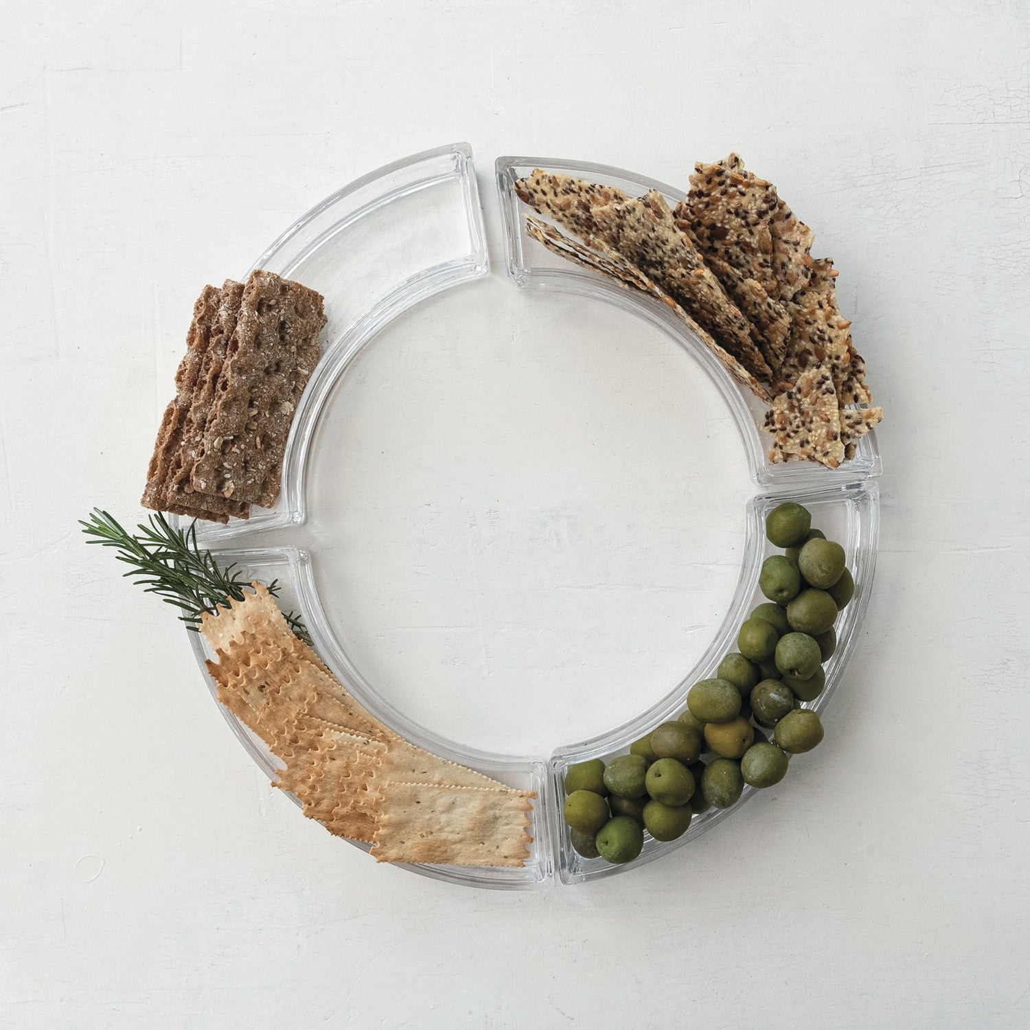 Circular clear serving tray containing crackers and green olives on a white surface