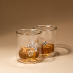 Studio product photograph featuring two double-walled glass tumblers with golden-amber whiskey and clear ice cubes on a light beige surface