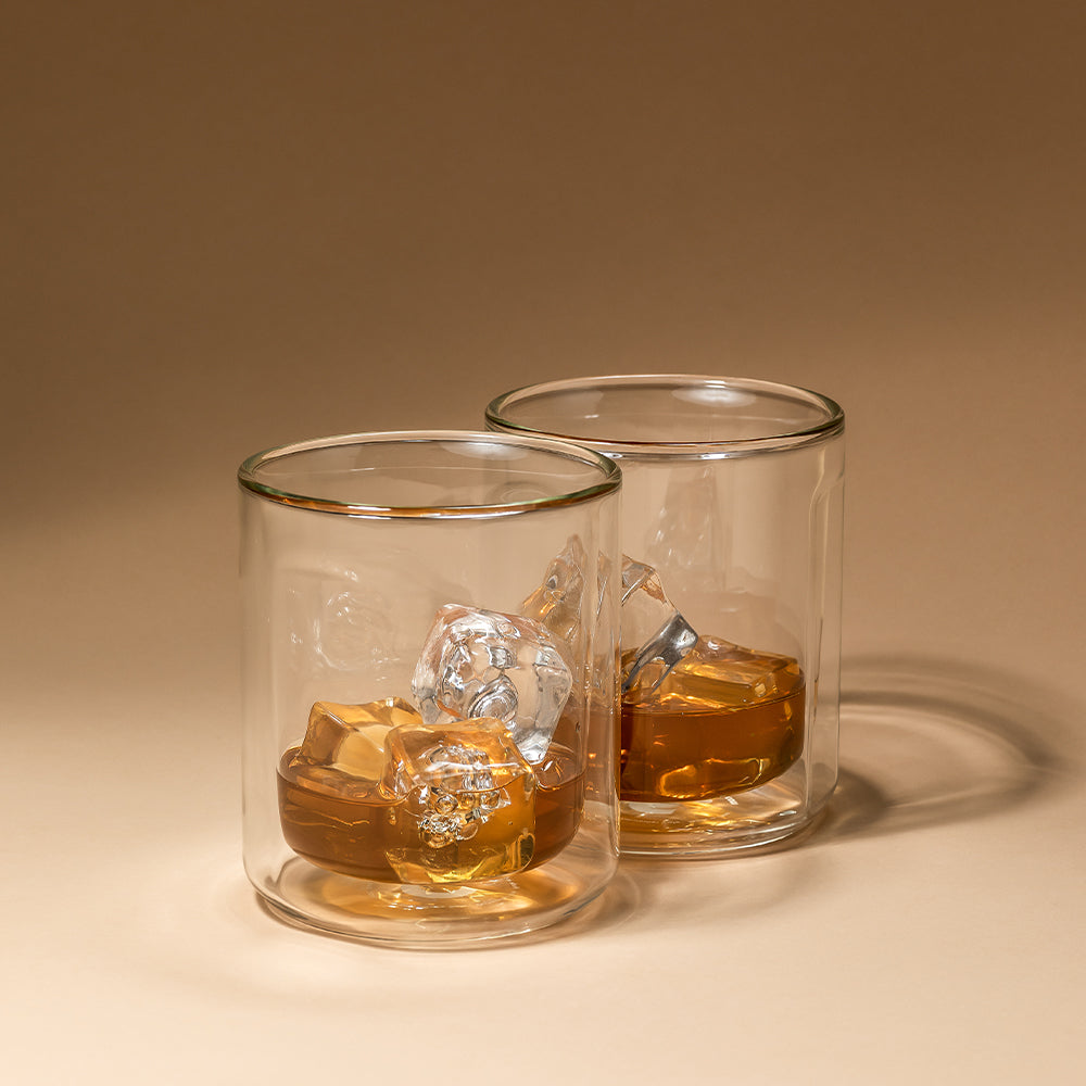 Studio product photograph featuring two double-walled glass tumblers with golden-amber whiskey and clear ice cubes on a light beige surface