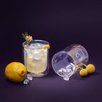 Product photography featuring two modern double-walled glasses on a deep purple background one containing an icy lemon cocktail garnished with rosemary and yellow flowers, the other iridescent and lying on its side displaying a C logo and ice, accompanied by a whole lemon and two yellow craspedia blooms