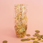 Photograph of a sparkling double-walled stemless champagne flute with gold 'cheers' text, filled with gold and iridescent hexagonal glitter, on a pale pink background scattered with gold circular confetti
