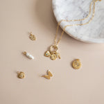 Studio photography of a gold paperclip chain necklace and assorted jewelry charms on a beige surface next to a white marble bowl featuring a white enamel pill charm with the word love, a crystal-accented palm tree, and a gold butterfly silhouette
