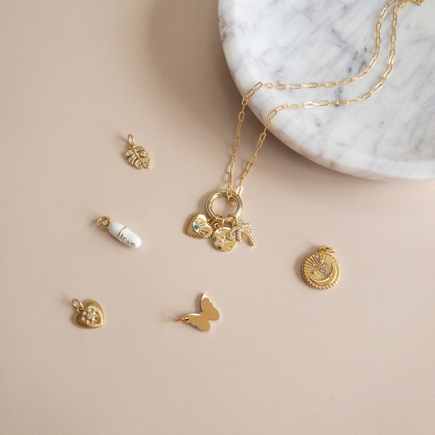Studio photography of a gold paperclip chain necklace and assorted jewelry charms on a beige surface next to a white marble bowl featuring a white enamel pill charm with the word love, a crystal-accented palm tree, and a gold butterfly silhouette