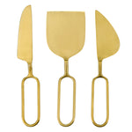 A modern minimalist brass set of three cheese tools featuring a knife, spreader, and cleaver with elongated oval open handles against a white background