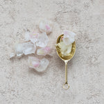 Gold spoon holding ice cubes with flower petals inside next to scattered ice cubes on a textured surface
