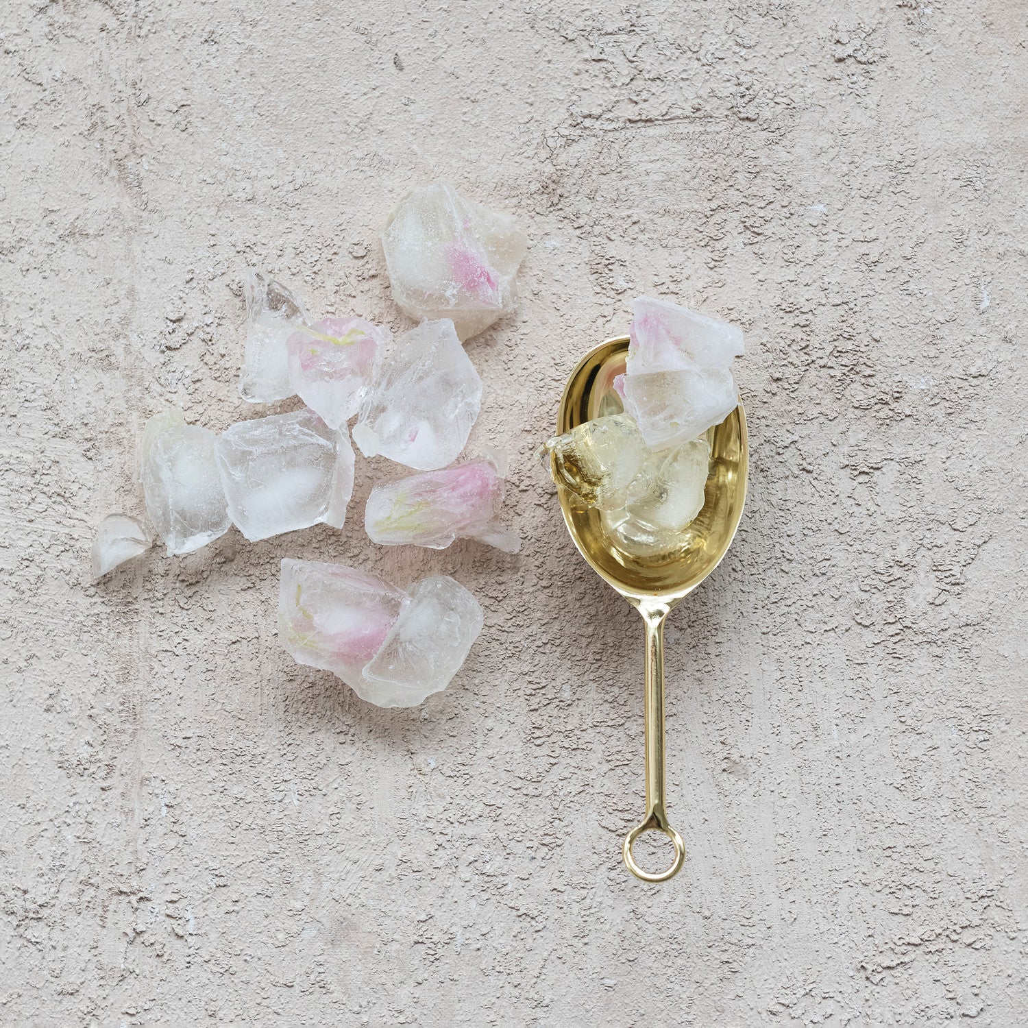 Gold spoon holding ice cubes with flower petals inside next to scattered ice cubes on a textured surface