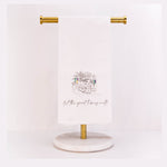 White linen towel featuring an illustrative sketch of a New Orleans street scene with a vintage streetcar and classic architecture, adorned with colorful Mardi Gras beads in purple, green, and gold, and the cursive phrase 'let the good times roll'