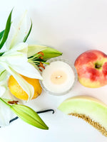 Lit candle surrounded by white lilies, an apple, a lemon, and a slice of melon