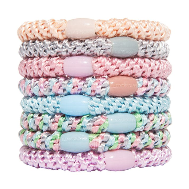 Digital photography by L. ERICKSON featuring a neatly stacked assortment of braided hair ties in various pastel shades including peach silver pink light blue mint green and lavender, each adorned with a branded oval bead