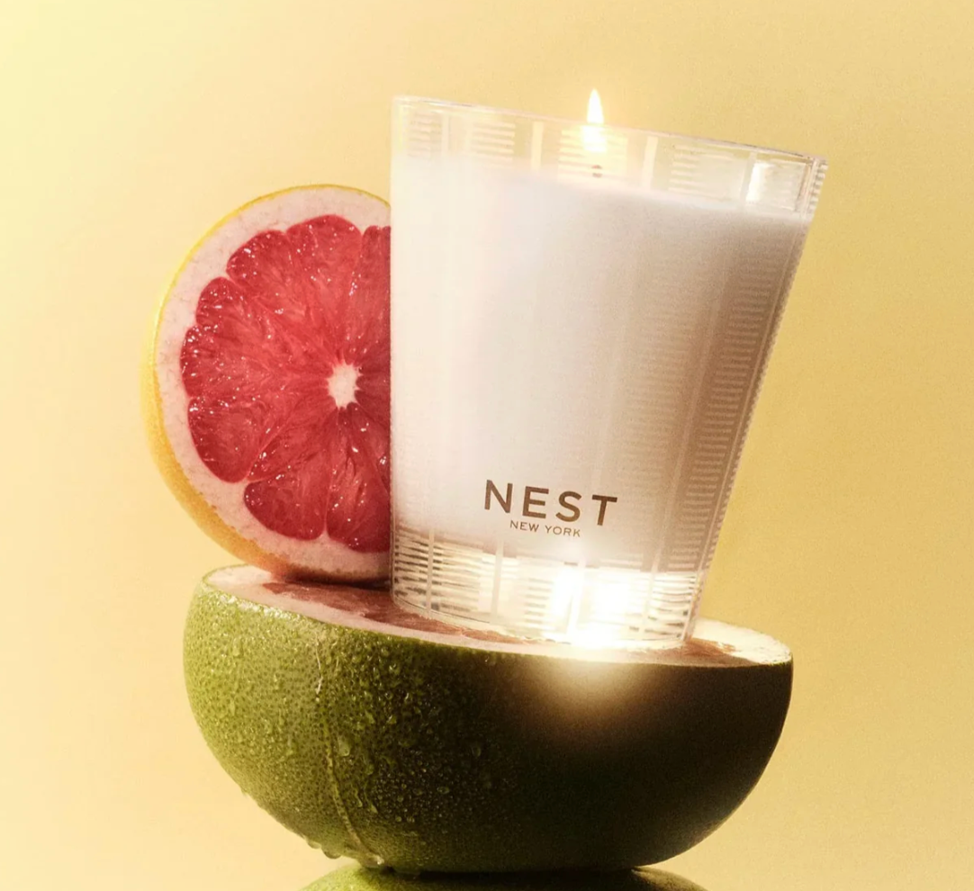 Product photography featuring a lit NEST New York grapefruit candle in a textured glass holder, positioned with a bright pink grapefruit half on a wet green citrus half, all against a sunny yellow background