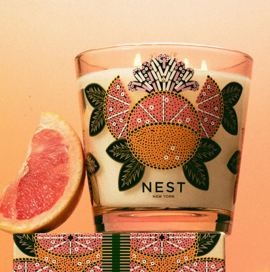 A photograph of a NEST New York three-wick grapefruit candle featuring a vibrant pink and orange stylized citrus pattern on its glass and matching box, presented next to a fresh pink grapefruit slice